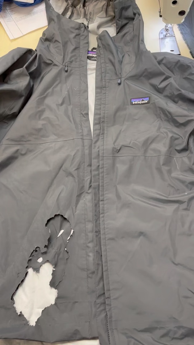 A gray Patagonia rain shell on a repair bench next to a sewing machine, a fist-sized hole torn through the front panel