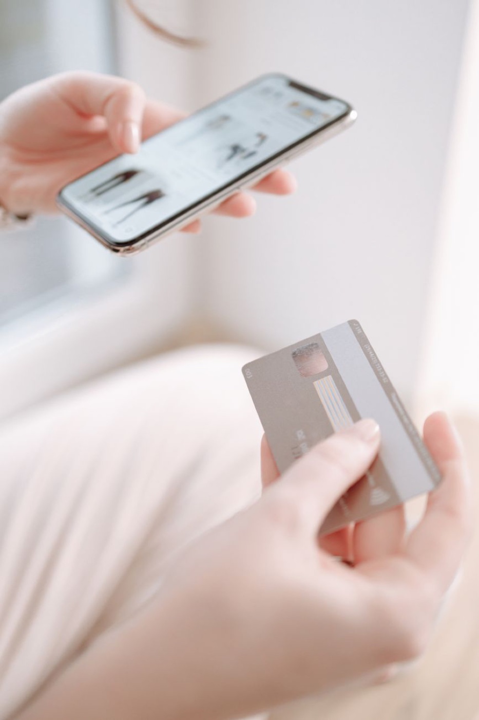 Hands holding a smartphone showing a clothing shopping app and a bank card by a window