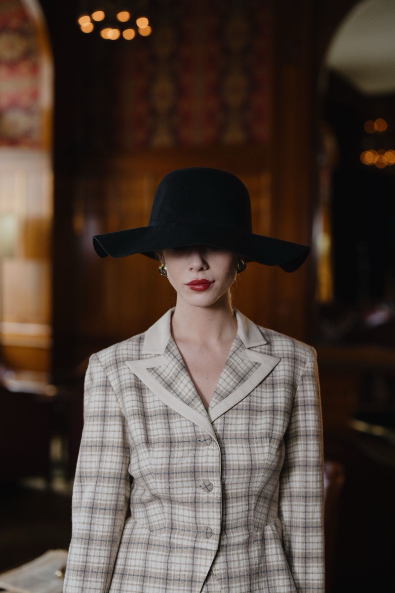 A woman in a beige check blazer and a wide-brimmed black felt hat that covers her eyes