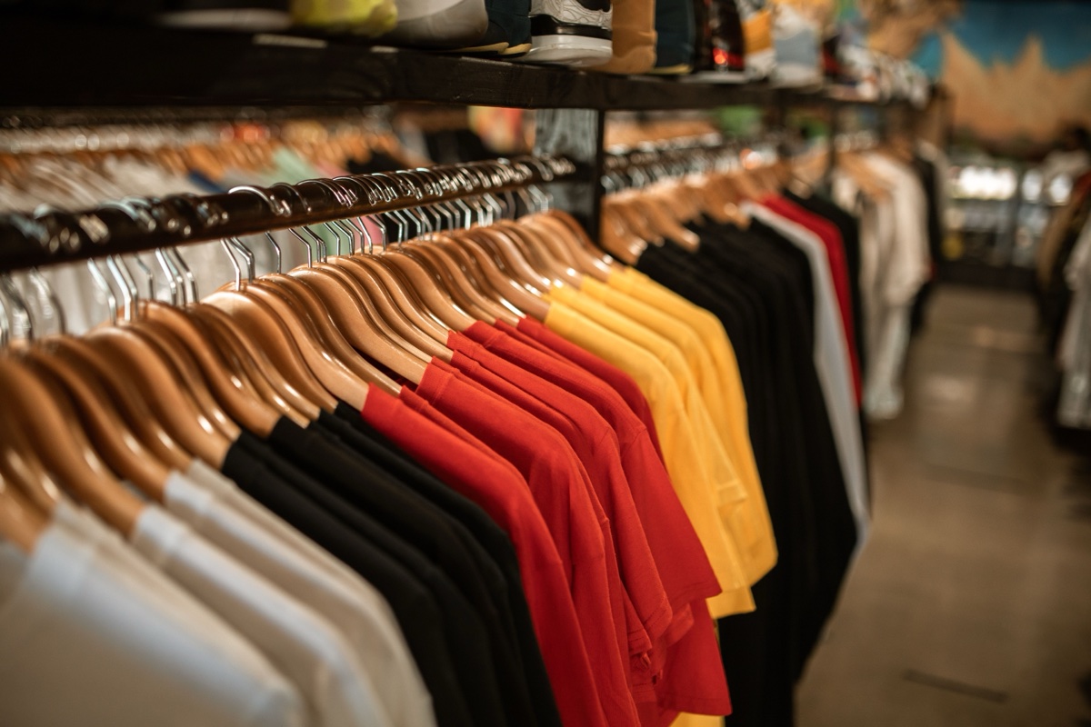 A long clothing rack of plain t-shirts in white, black, red, and yellow on wooden hangers with sneakers on a shelf above