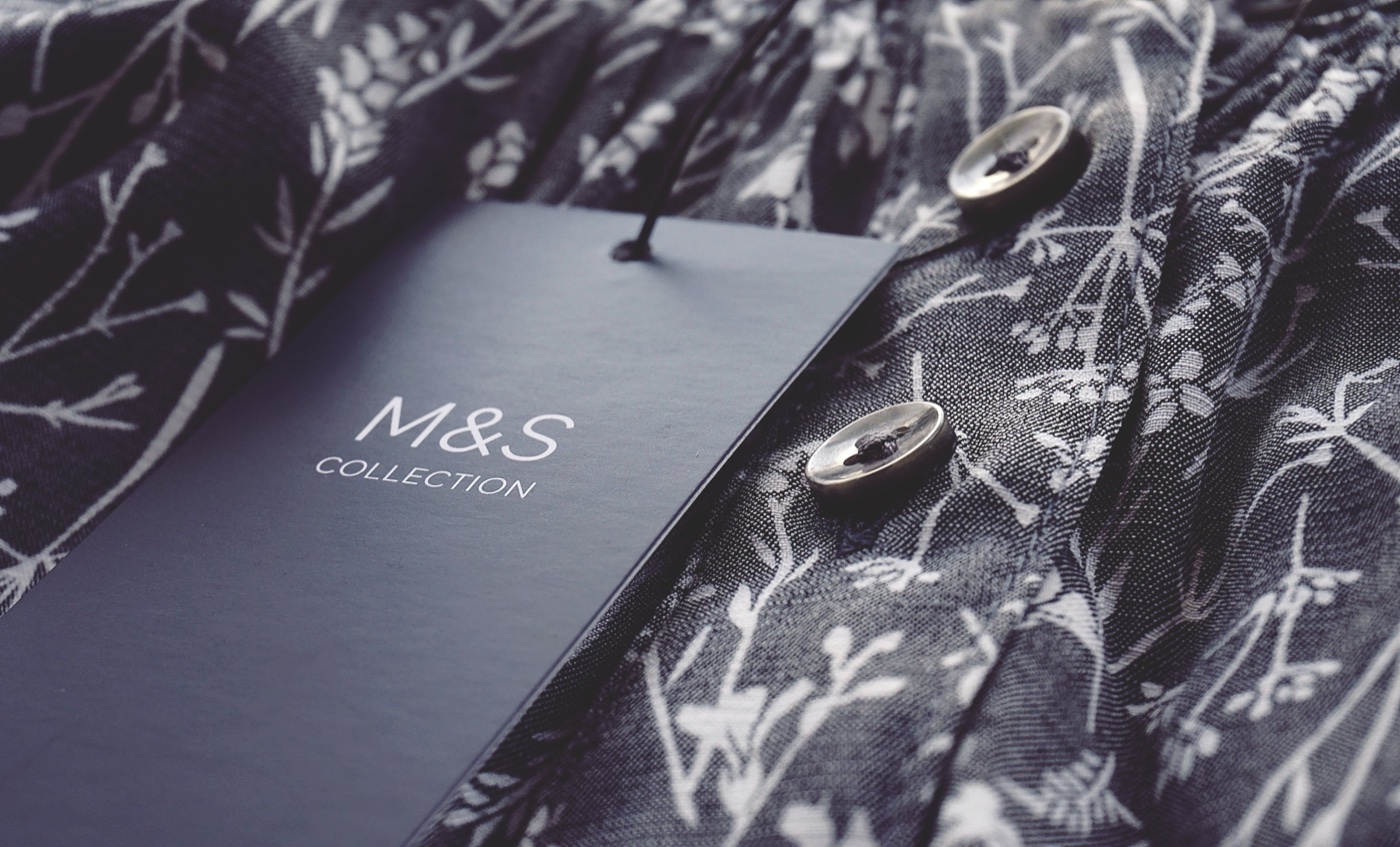 A navy M&S Collection hang tag resting on a dark floral-print blouse with two metal buttons, shot close enough to show the weave of the fabric