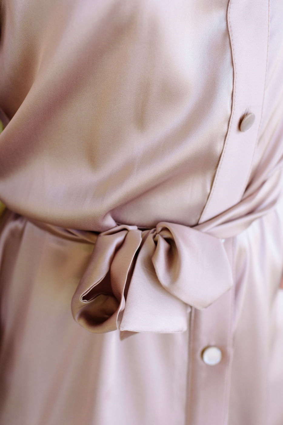 Close-up of a blush pink satin shirtdress with a self-tie bow at the waist and two fabric-covered buttons, the fabric catching soft light