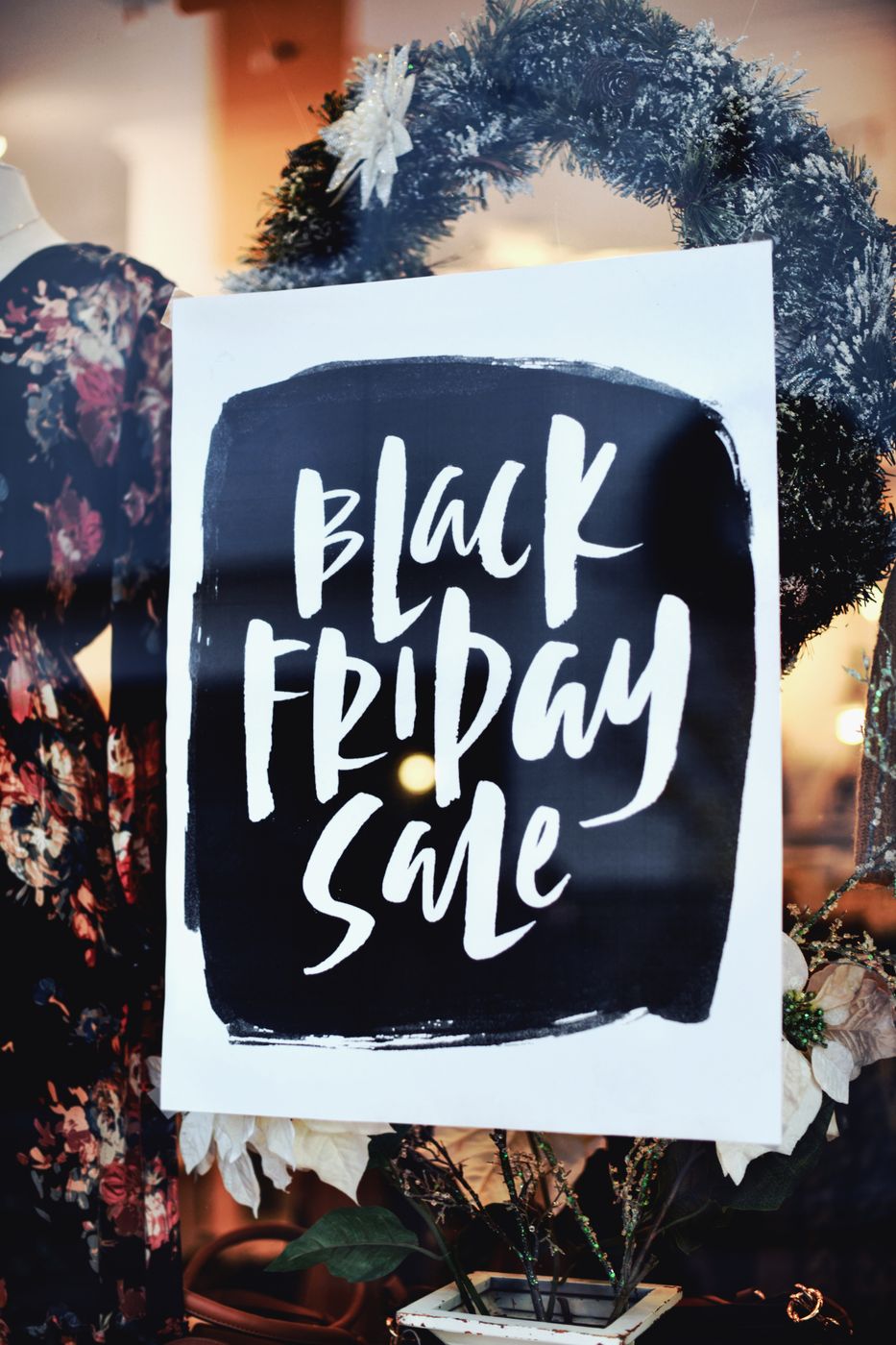 A hand lettered Black Friday Sale poster in a boutique window, with a floral dress and a wreath visible behind the glass