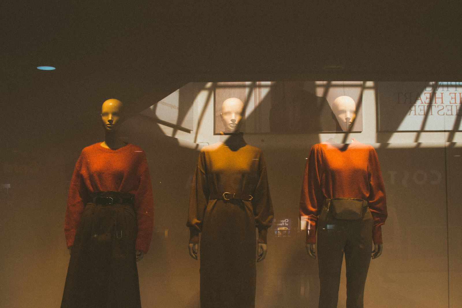 Three mannequins in rust and olive knits standing behind a shop window in low amber light
