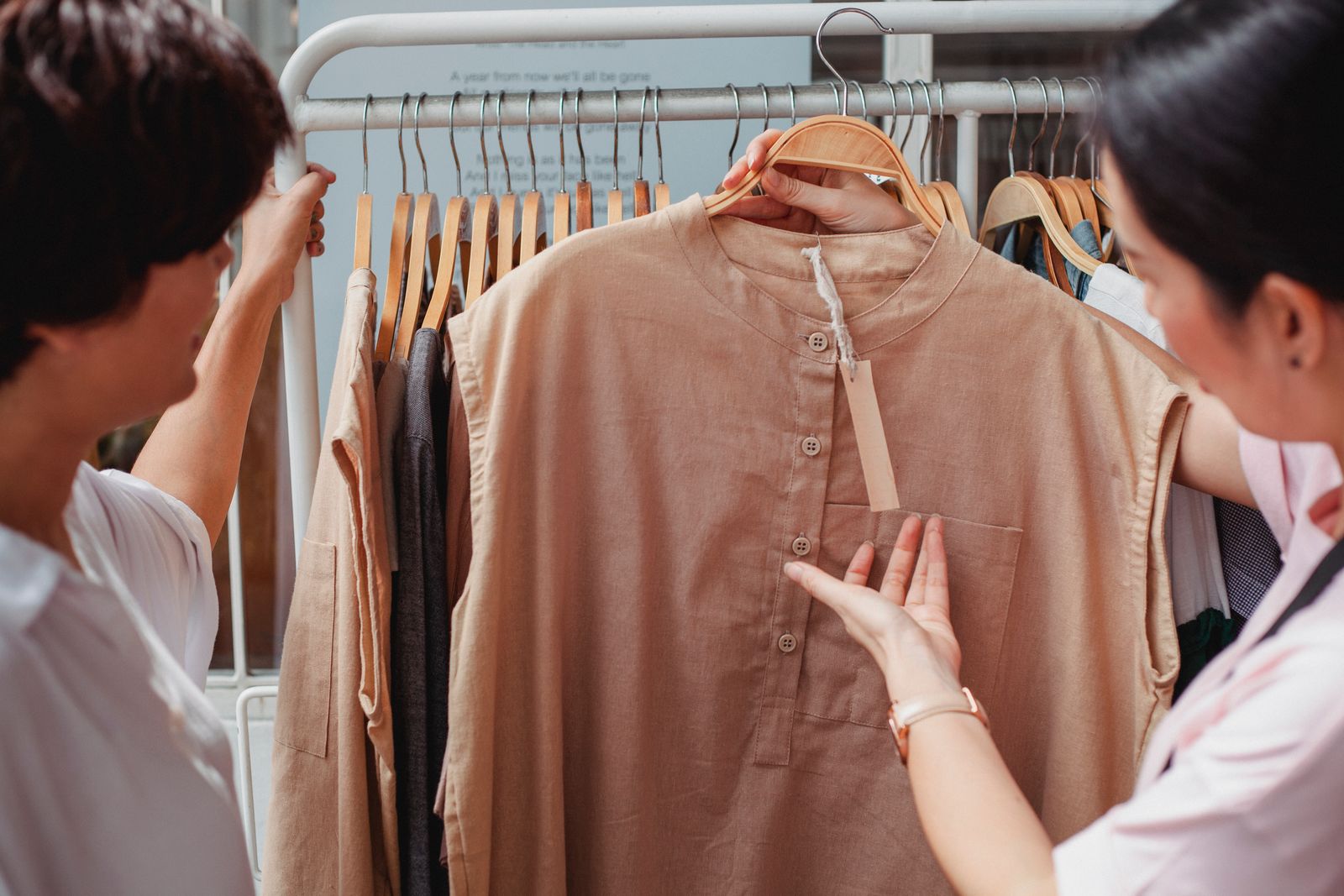 Two shoppers at a rack of tan shirts, one holding out a sleeve to read the blank hang tag at the collar