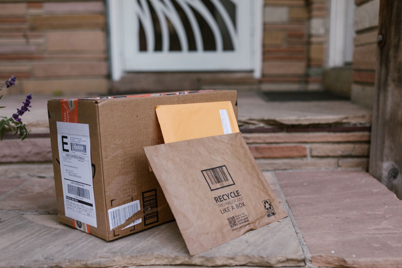 Cardboard shipping box and paper mailers stacked on a front doorstep beside a purple flower