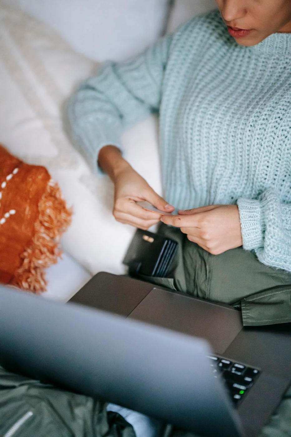 Overhead view of a woman in a mint knit sweater holding a bank card beside an open laptop on a bed