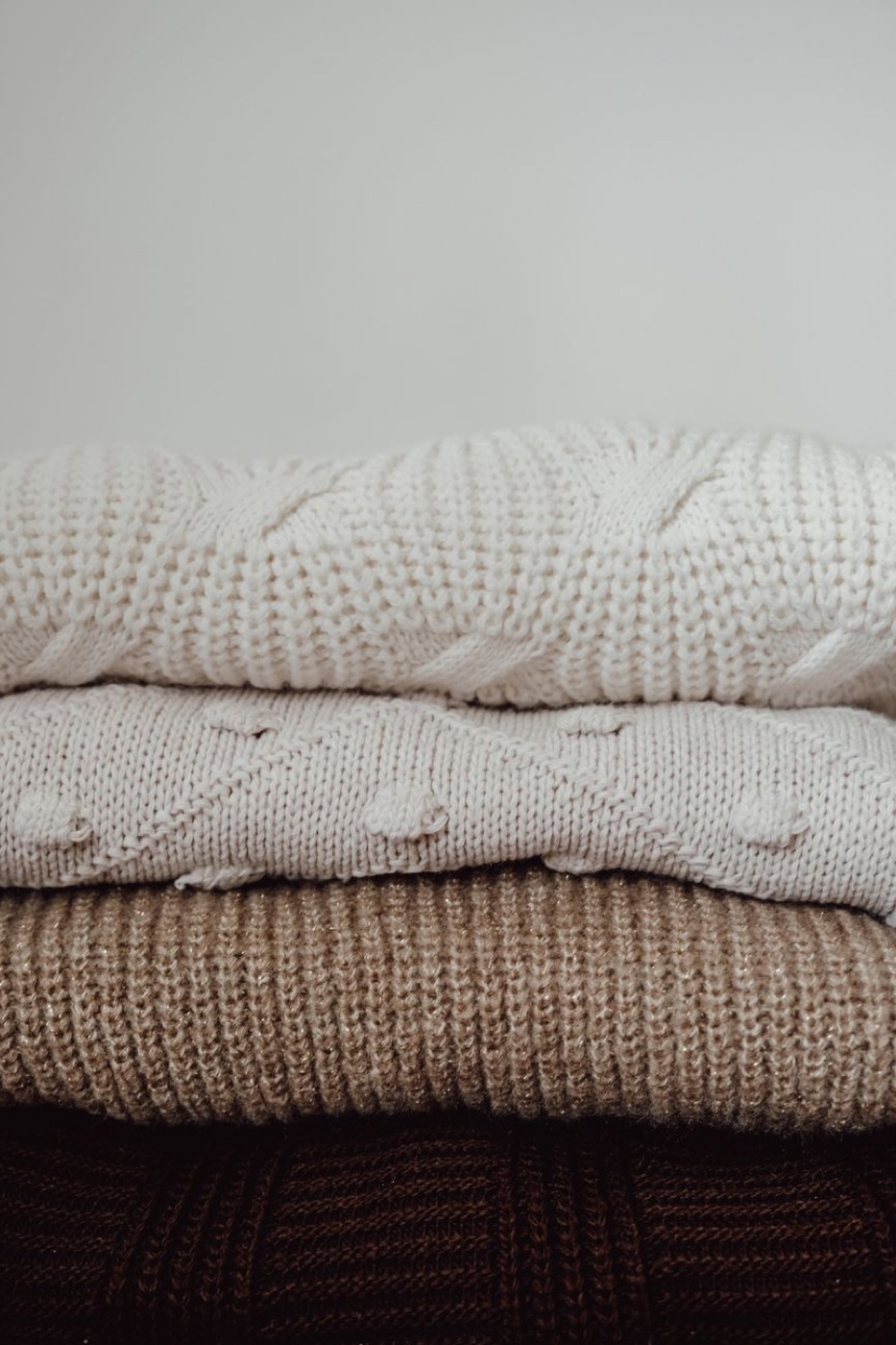 Stack of folded cream, beige and brown knit sweaters against a plain wall