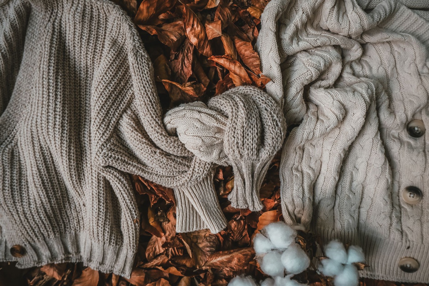 Two beige cable-knit sweaters and a cardigan laid over dry autumn leaves