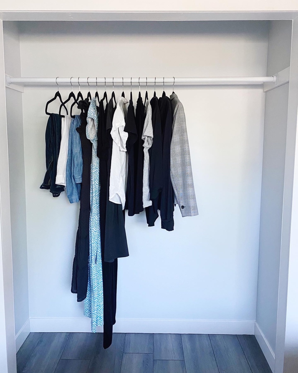 A closet with a single rail holding about twenty hangers of tops, shirts, trousers and a blazer, the floor bare beneath them