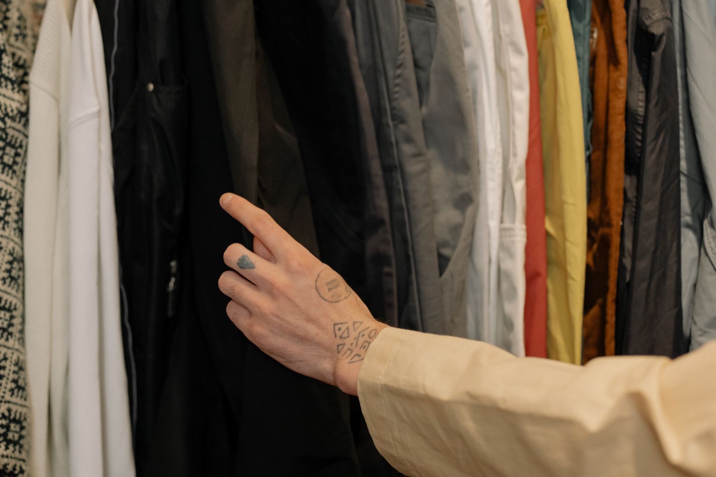A tattooed hand reaching into a densely packed rail of jackets and shirts to pull one forward