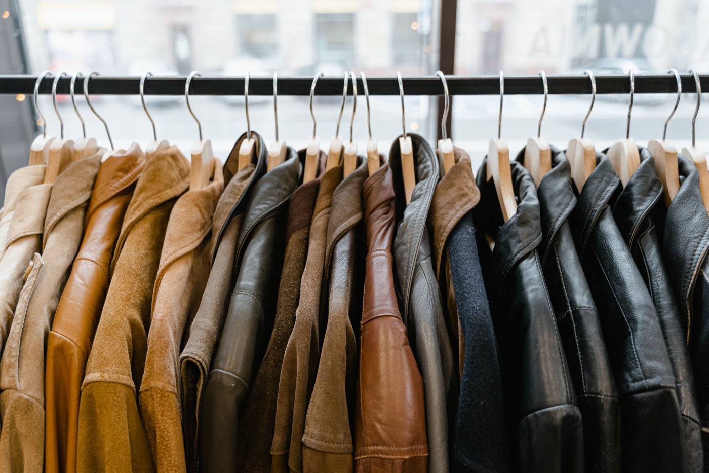 A dense rail of leather and suede jackets in tan, brown, grey and black on wooden hangers in front of a shop window