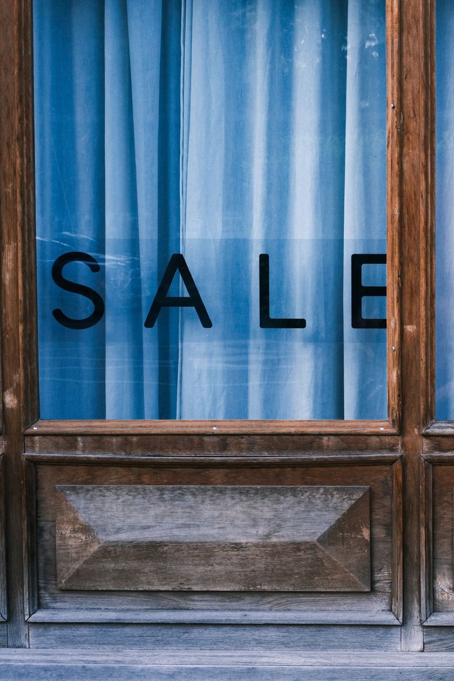 Black SALE lettering applied to the glass of a wooden shopfront with a blue curtain drawn behind it