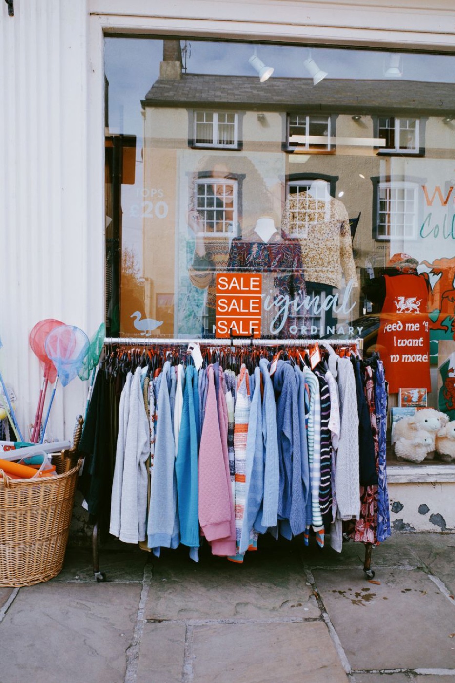 A wheeled rail of knitwear parked on a pavement outside a shop window with three orange SALE cards clipped above it