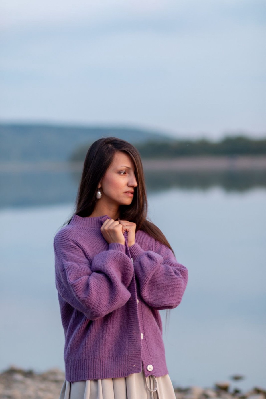 Woman in profile by a still lake at dusk, holding closed the collar of a plum purple button-up cardigan