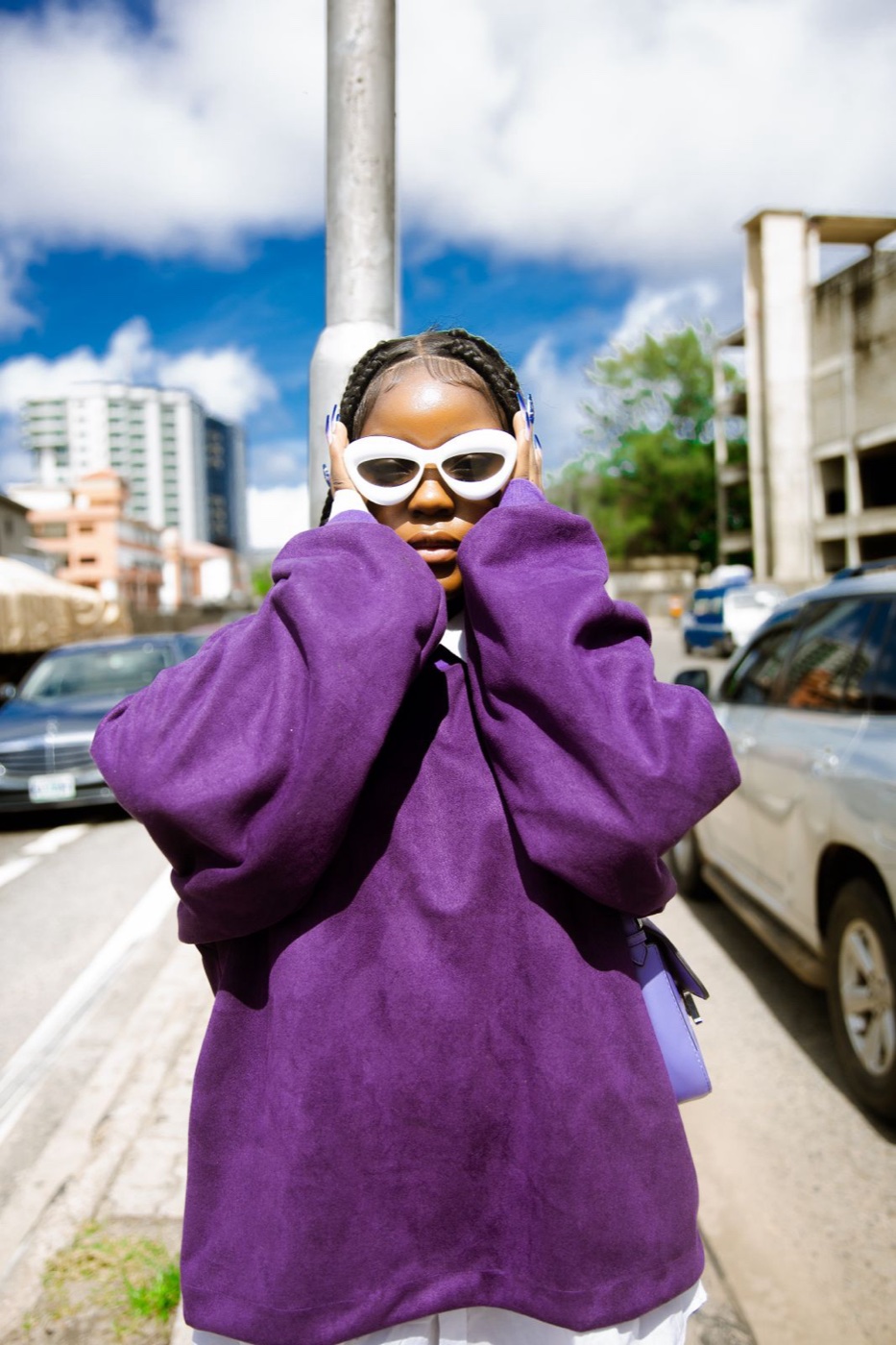Woman on a city sidewalk in an oversized deep purple sweatshirt, holding white sunglasses to her face, parked cars behind her
