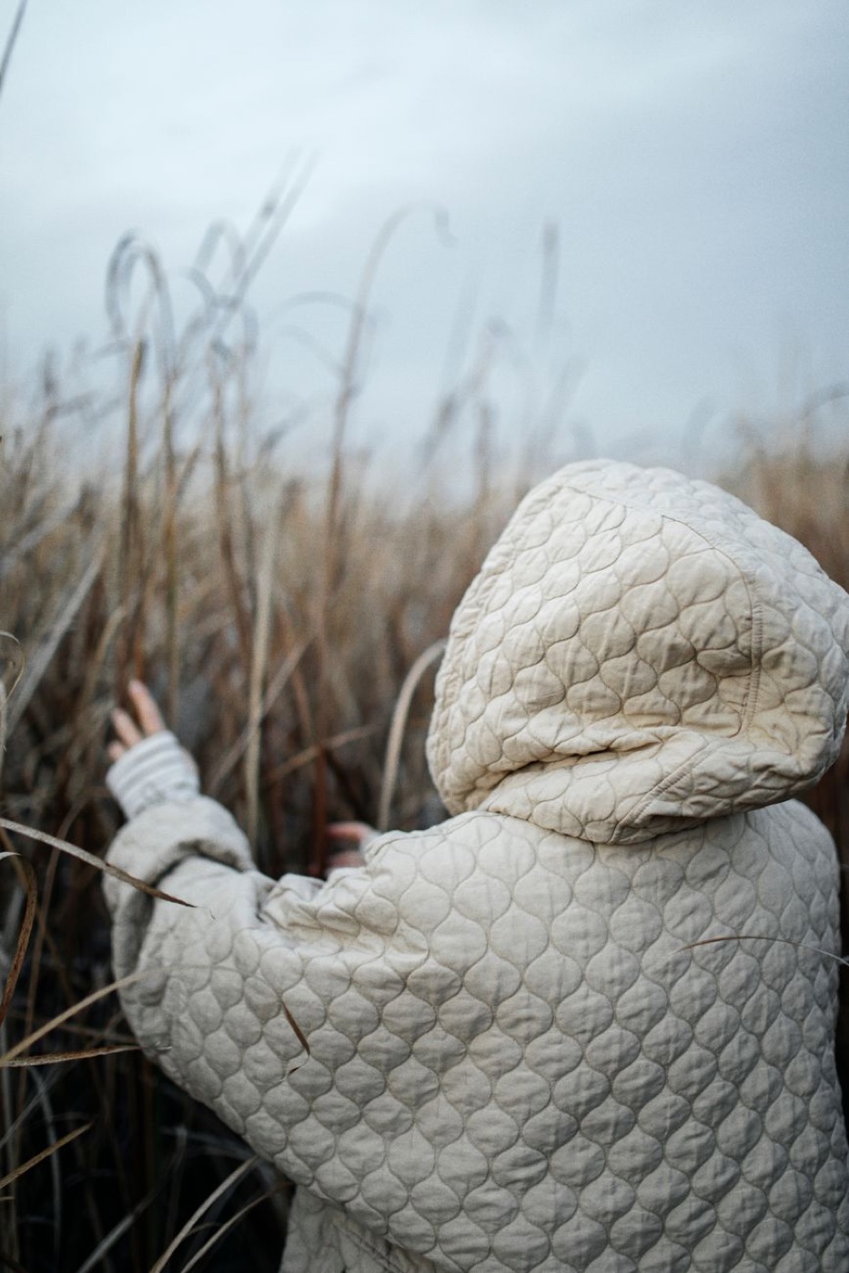 Rear view of a person in a cream quilted hooded jacket standing in tall dry grass