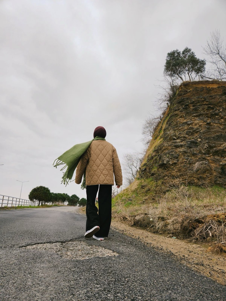 Person seen from behind in a quilted tan jacket and green scarf walking along an empty country road under an overcast sky