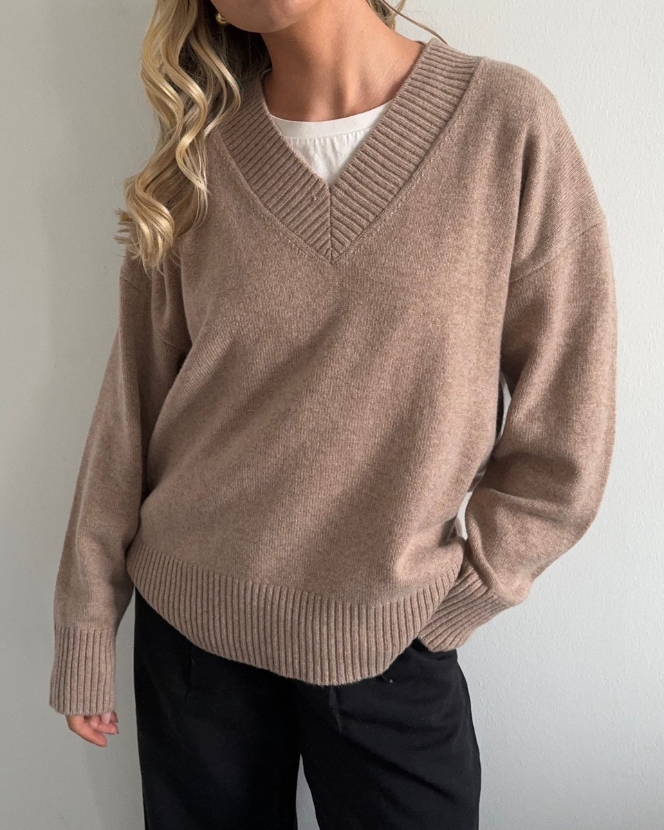 A camel cashmere V-neck sweater worn over a white tee with black trousers, ribbed cuffs and hem, shot against a plain wall