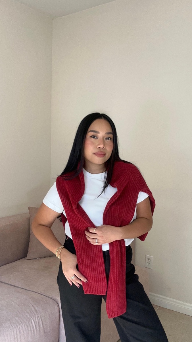 A woman with a red waffle-knit cashmere sweater draped over a white tee, standing beside a mauve sofa