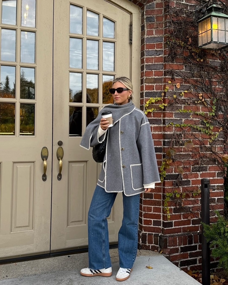 A woman in a grey scarf coat with cream whipstitch trim holds a coffee outside a paneled door, wearing jeans and white sneakers