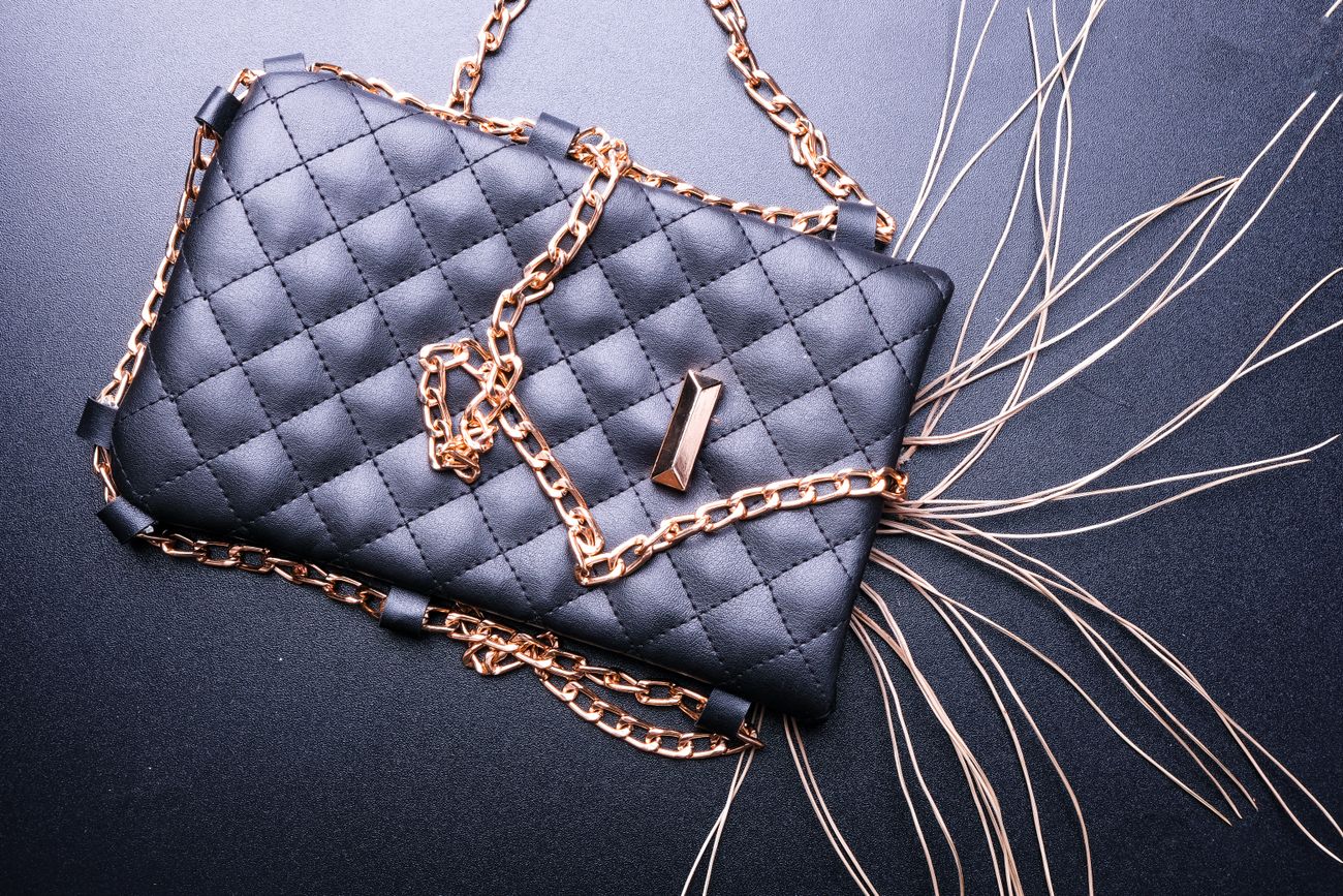 A black quilted crossbody bag with a gold chain strap laid flat on a dark surface beside pale fringe trim