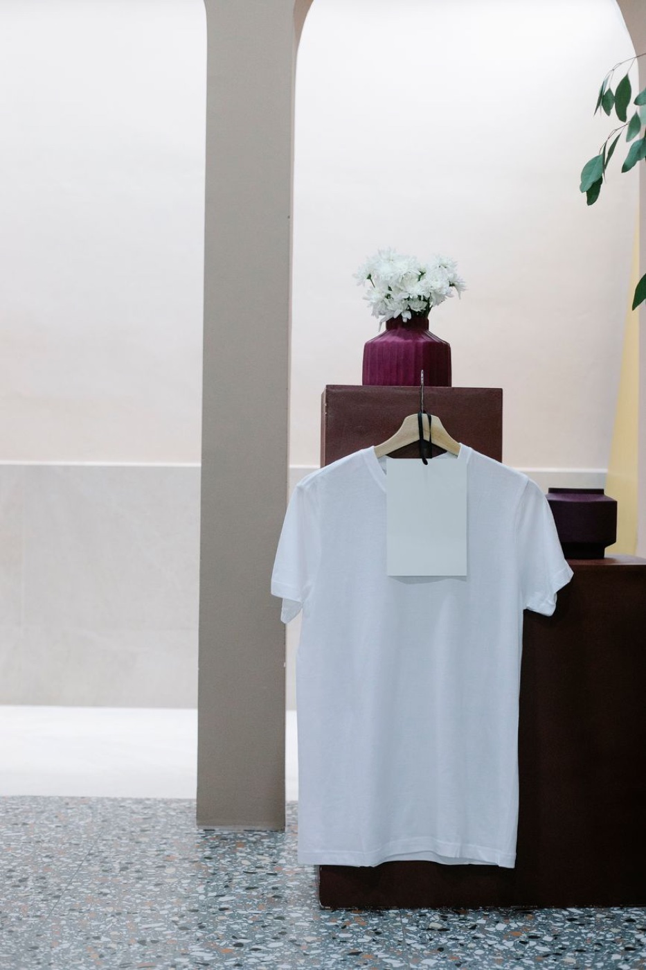 A plain white t-shirt on a wooden hanger in a pale shop interior, with a large blank paper swing tag hanging over the chest