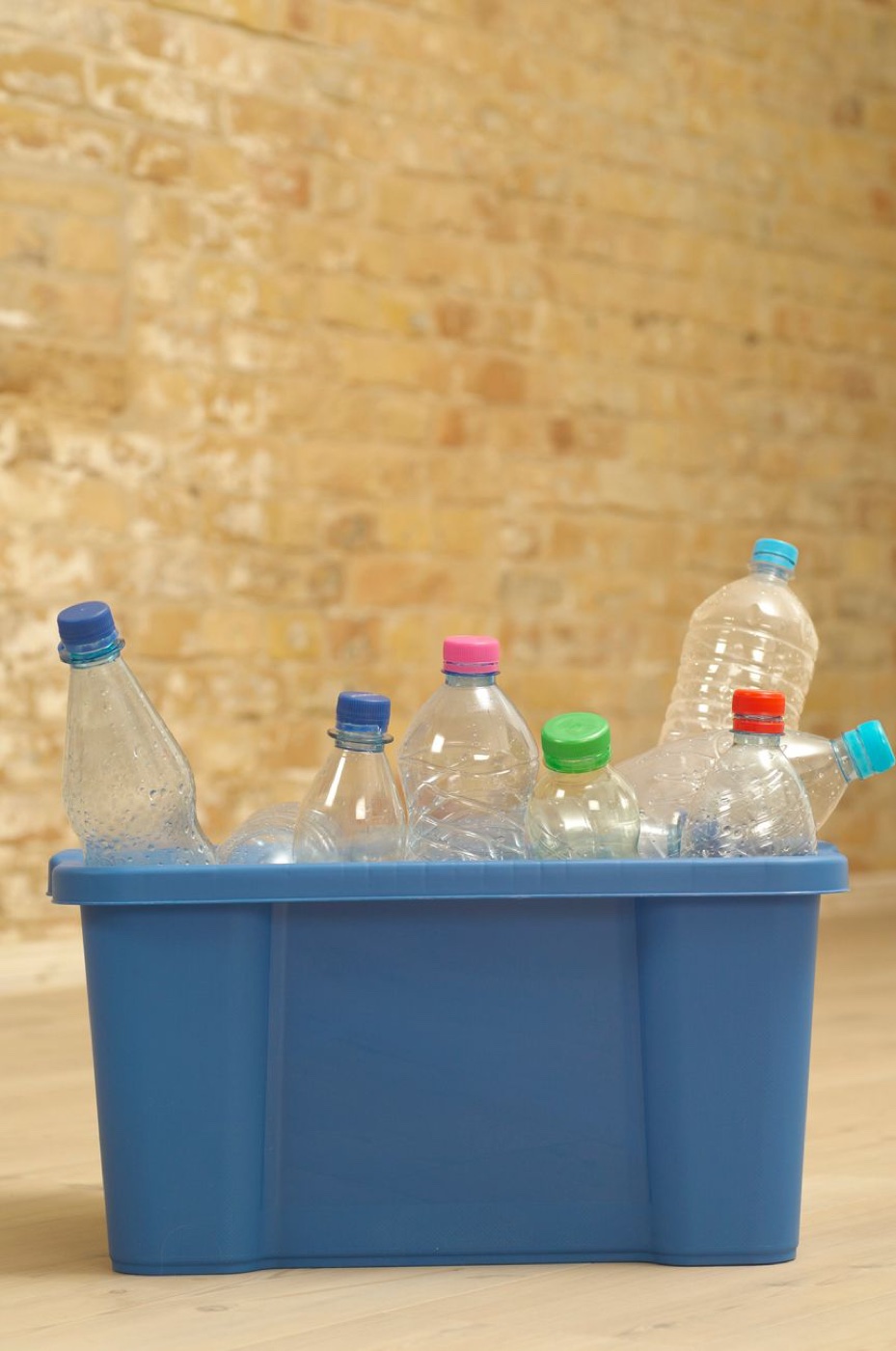 Seven clear plastic drinks bottles with coloured caps standing in a blue kerbside recycling box against a brick wall
