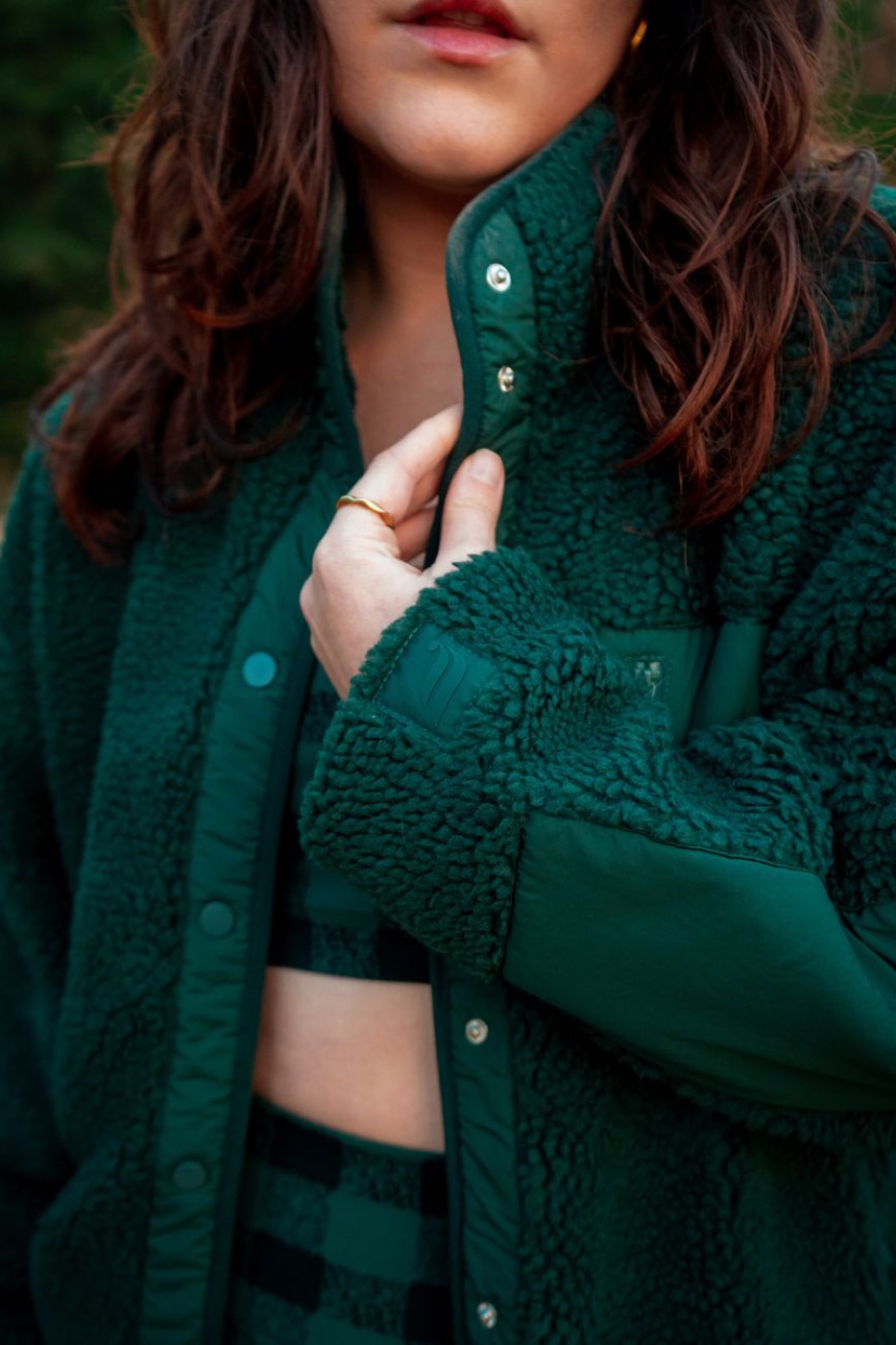 A person in a dark green sherpa fleece jacket with a nylon yoke, holding the collar closed, photographed outdoors from the shoulders down