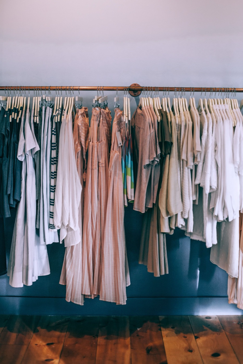 A copper rail against a pale wall holding rows of pale dresses, striped shirts and knits on wooden hangers above a wood floor