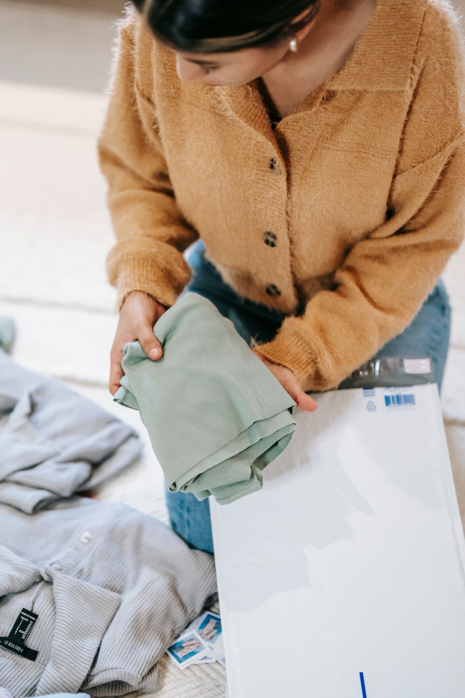 A person in a mustard cardigan kneeling on a rug, folding a sage green ribbed knit above an open white poly mailer with more clothes beside them