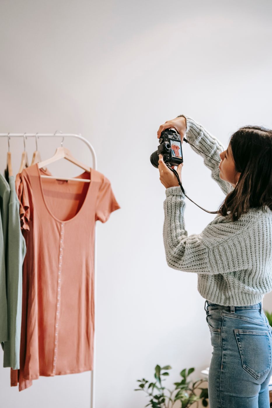 Woman in a pale knit sweater photographing a rust-coloured dress hanging on a white clothing rail