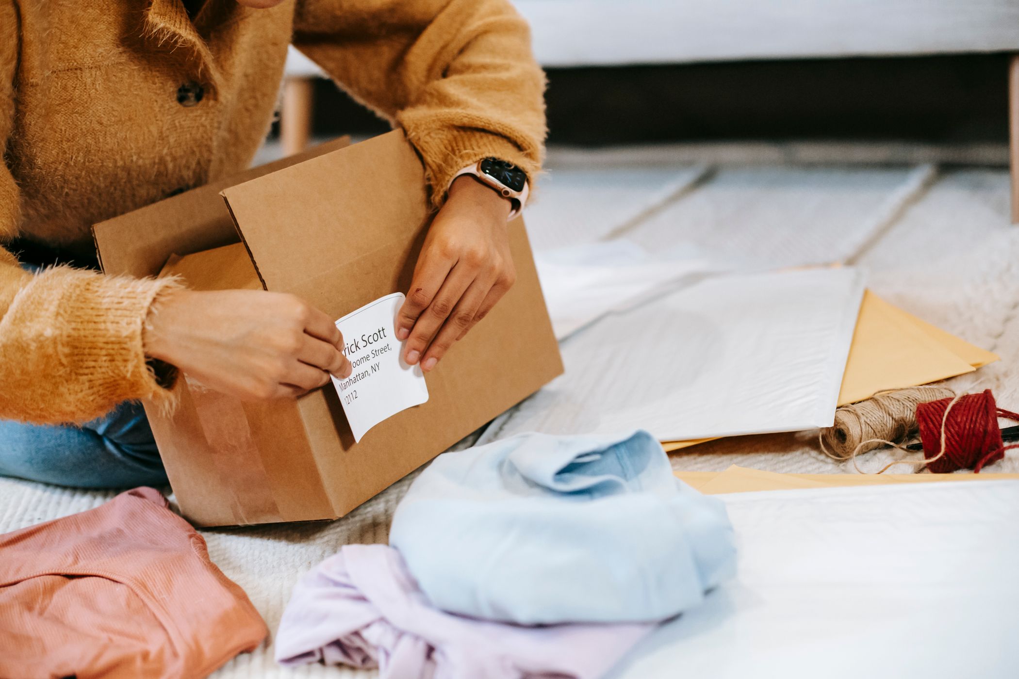 Hands in a mustard sweater pressing a printed address label reading Manhattan NY onto a cardboard box, folded clothes nearby