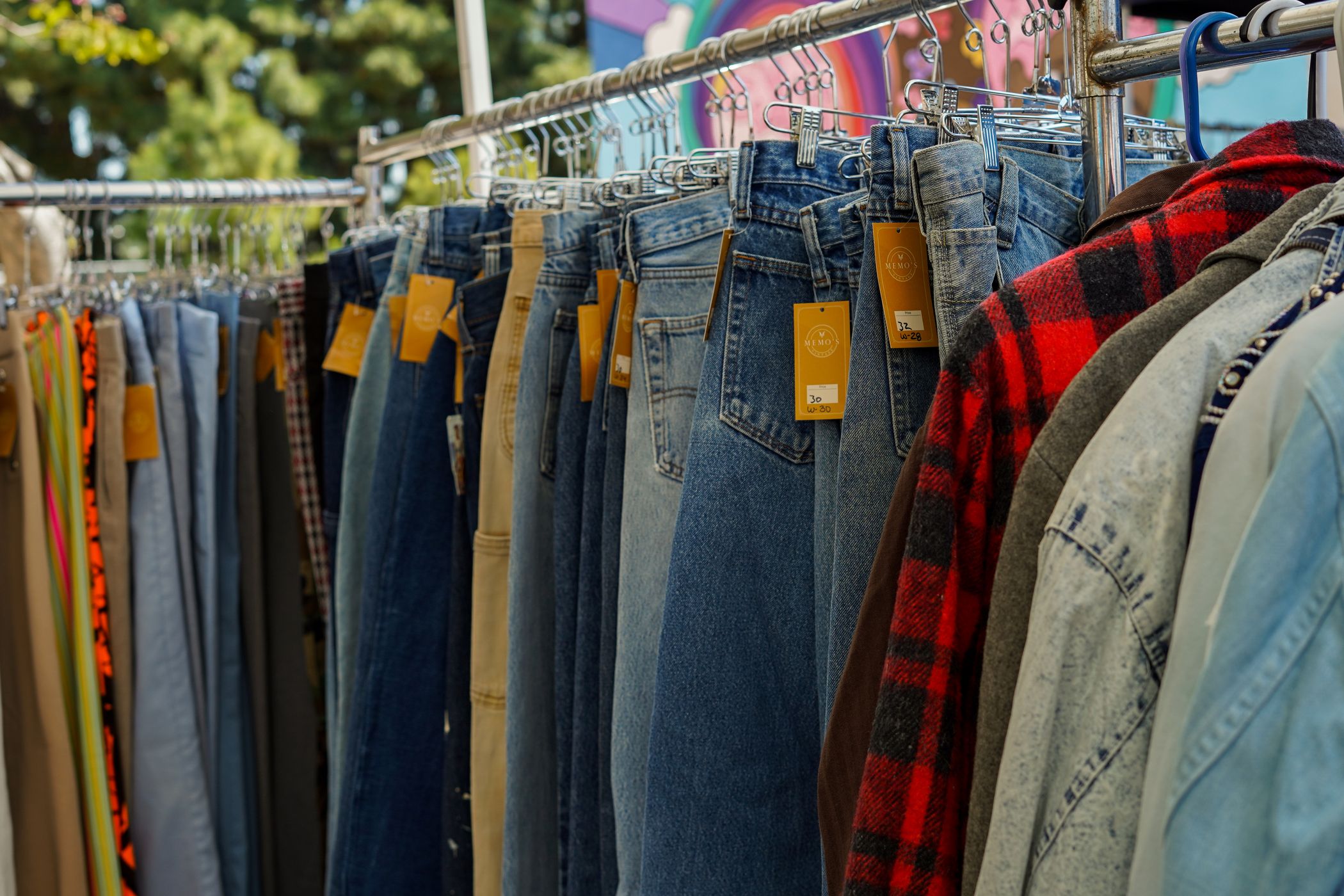 Rack of vintage jeans and jackets at an outdoor market with handwritten orange price tags reading 30 and 32