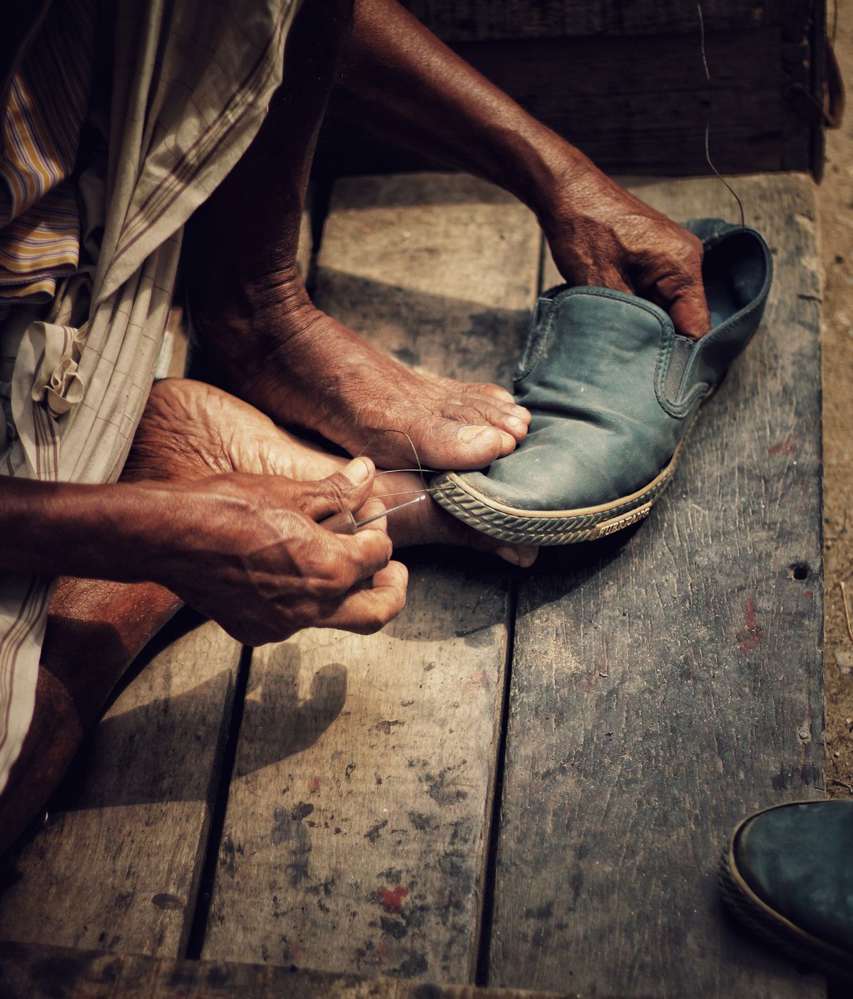 Two weathered hands drawing a waxed thread through the welt of a blue leather shoe on a wooden floor