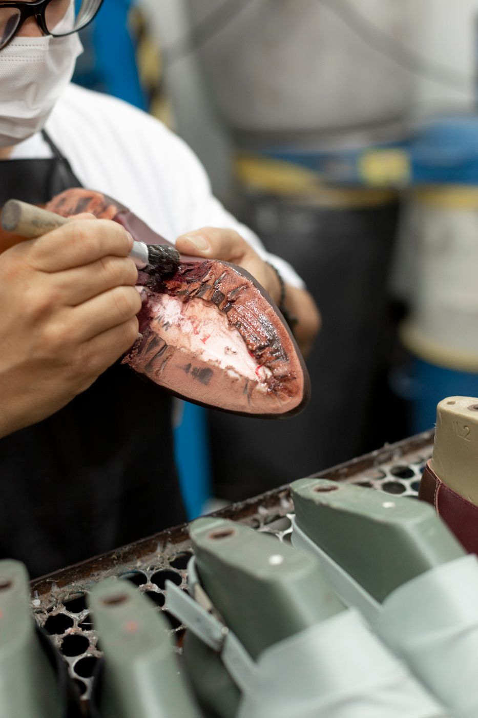 A masked worker in a shoe factory brushing adhesive onto the edge of a sole held over a tray of plastic lasts