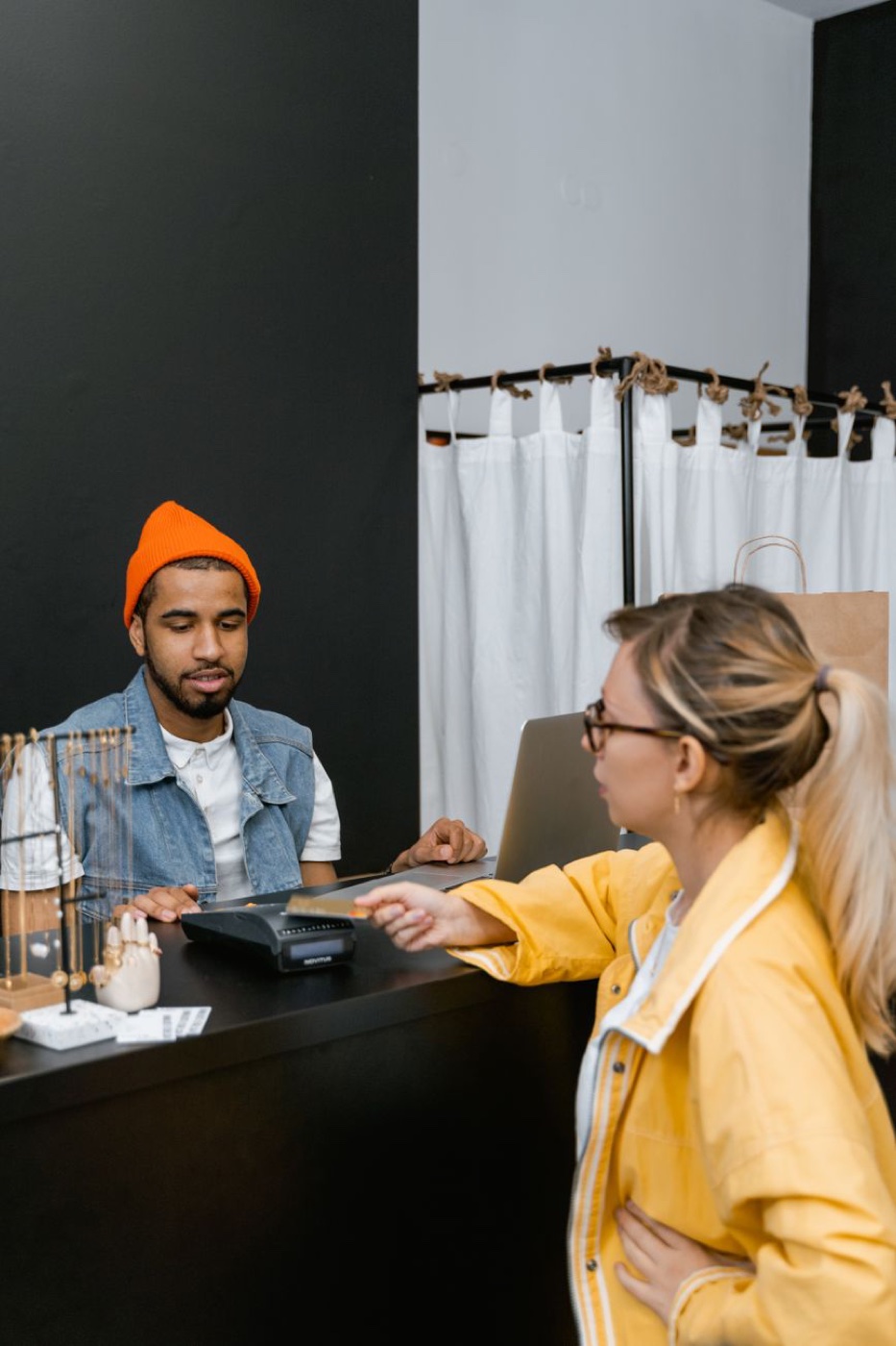 A shopper in a yellow jacket holding out a card to a payment terminal on a black counter while a shop assistant in an orange beanie watches