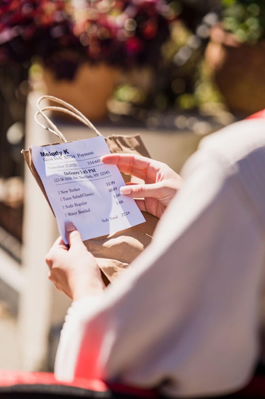 A person in white sitting outdoors reading an itemized paper receipt held against a brown takeaway bag in sunlight