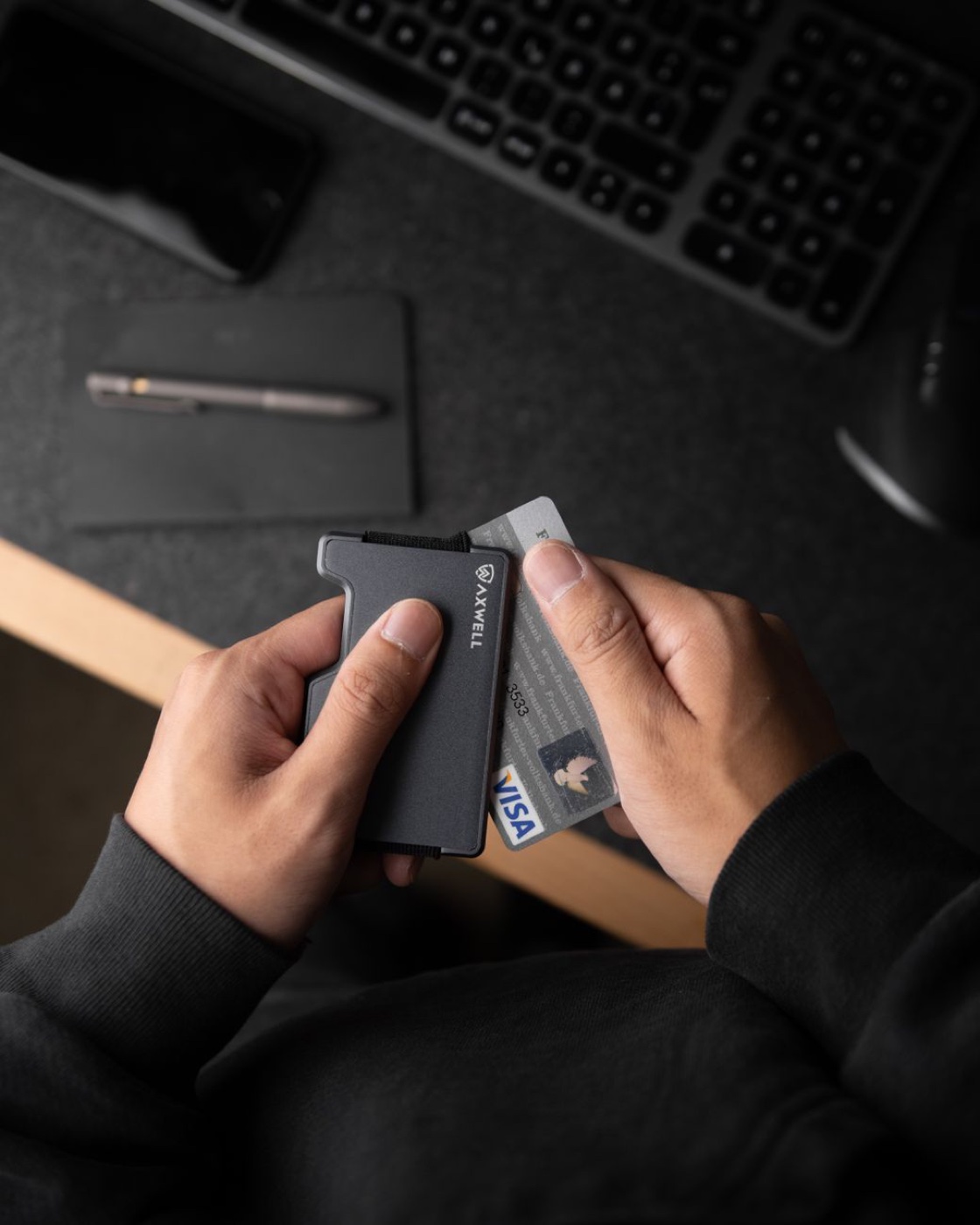 Two hands holding a slim metal wallet with a Visa card and a photo ID card sliding out of it above a dark desk