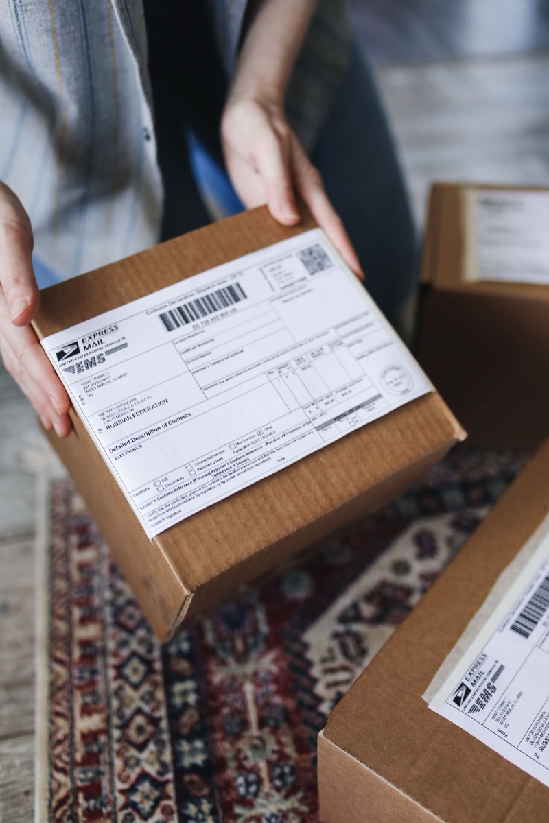 Hands holding a small cardboard box with a printed express mail label on top, more labeled boxes on the rug below
