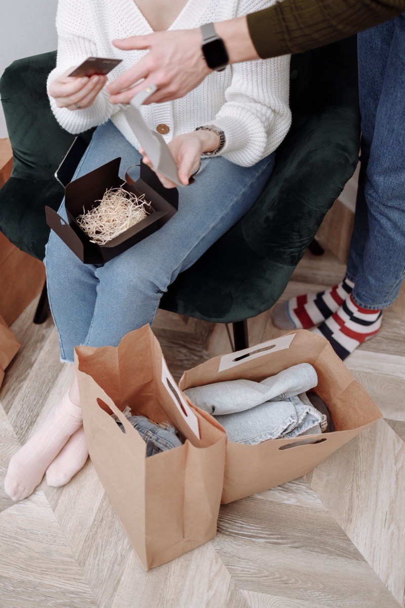 Two paper shopping bags of folded clothes on a herringbone floor beside a seated person holding a receipt and card