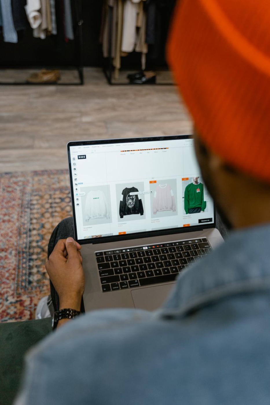 A person in an orange beanie looking at an online clothing store on a laptop while sitting inside a shop with racks behind them