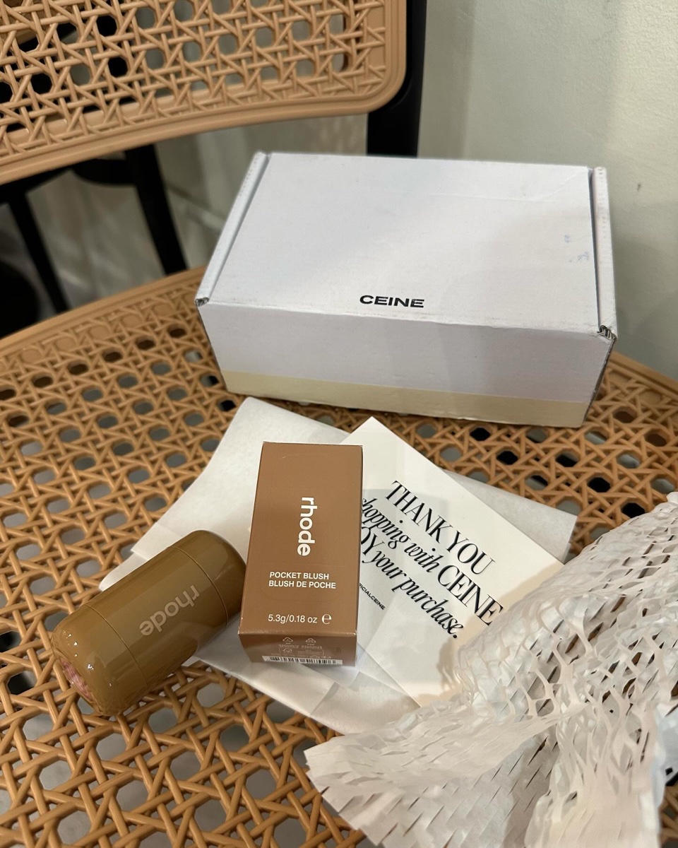 A Rhode Pocket Blush stick and its brown carton printed 5.3 g / 0.18 oz on a woven cane chair with shipping wrap