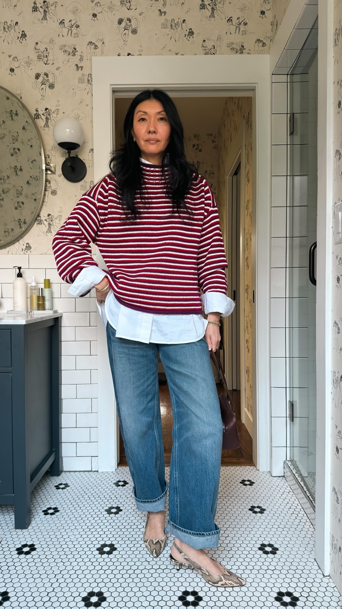 Woman standing in a tiled bathroom doorway wearing a red and white microstripe rollneck over a white poplin shirt, cuffed jeans and snake-print slingbacks