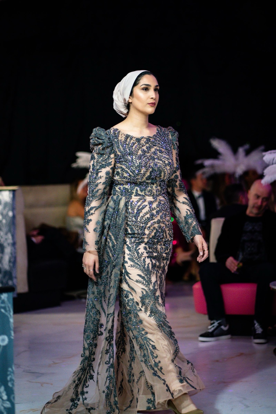 A model on a runway in an embroidered sheer gown and a white headscarf, with feathered headpieces visible in the front row behind her