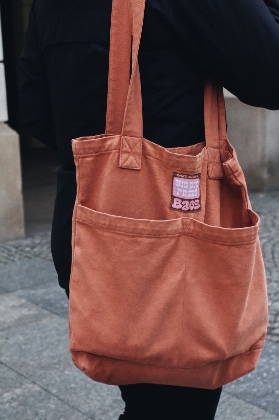 Close view of a rust-colored cotton twill tote bag with reinforced stitched straps carried over a shoulder on a cobblestone street