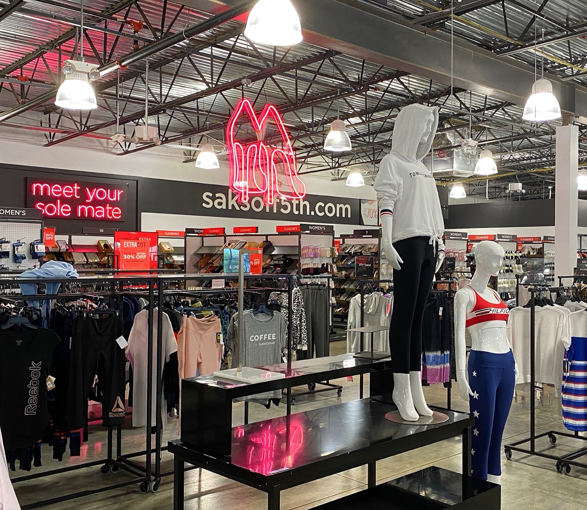 Interior of a Saks Off 5th store with a saksoff5th.com wall sign, a neon heels graphic, and clearance racks of activewear