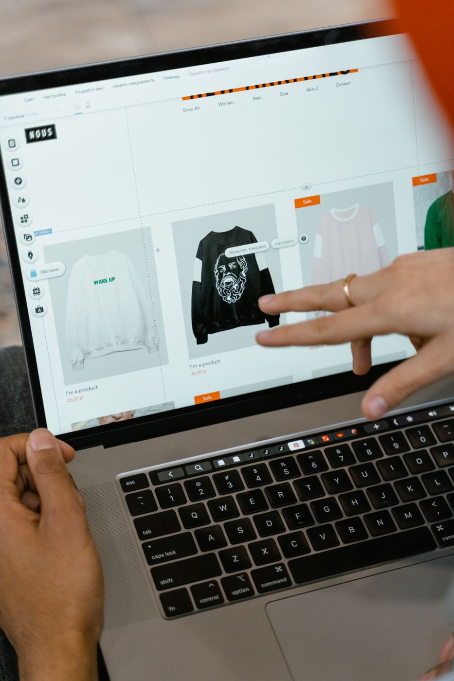 A hand pointing at a laptop screen showing an online clothing shop with orange sale badges on sweatshirt listings