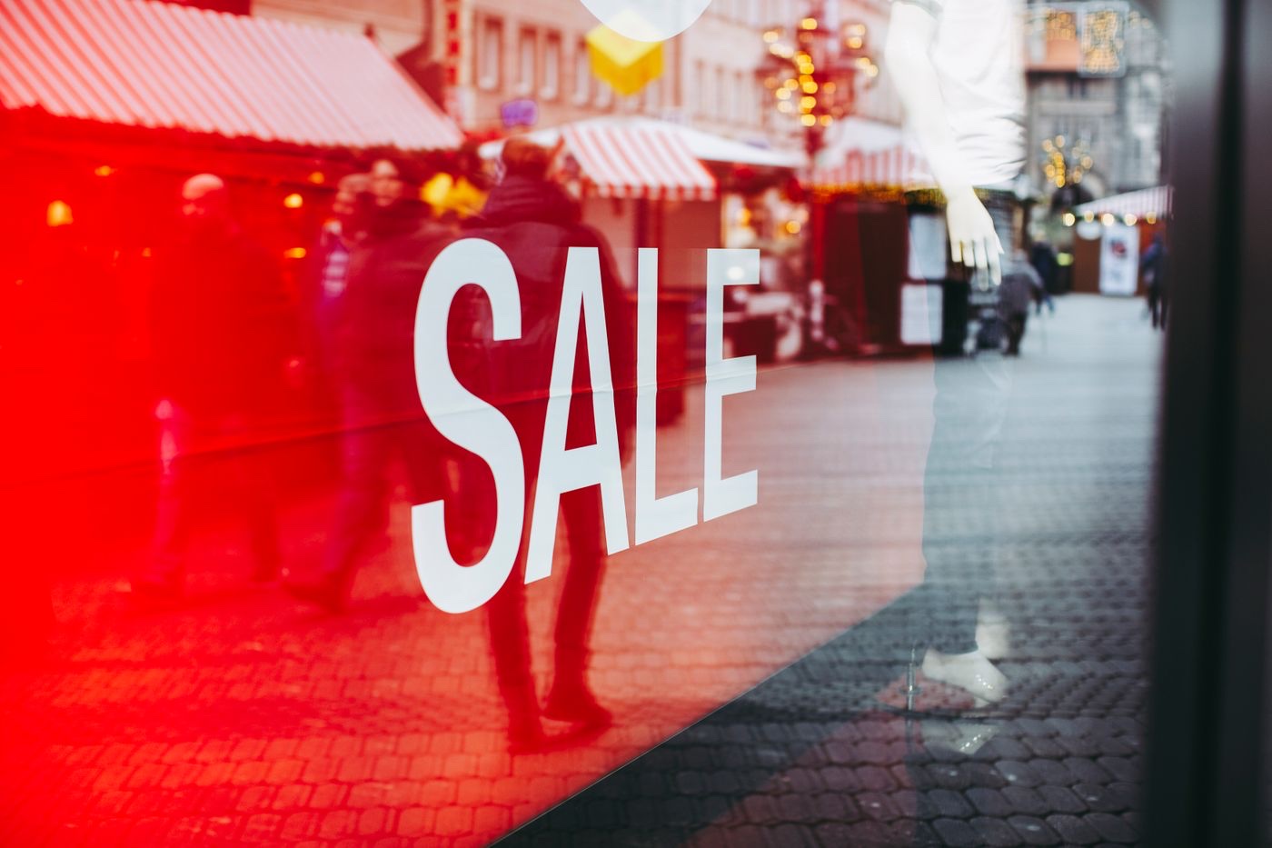 A large white SALE decal on a shop window, with a red interior display and blurred street reflections of shoppers across the glass