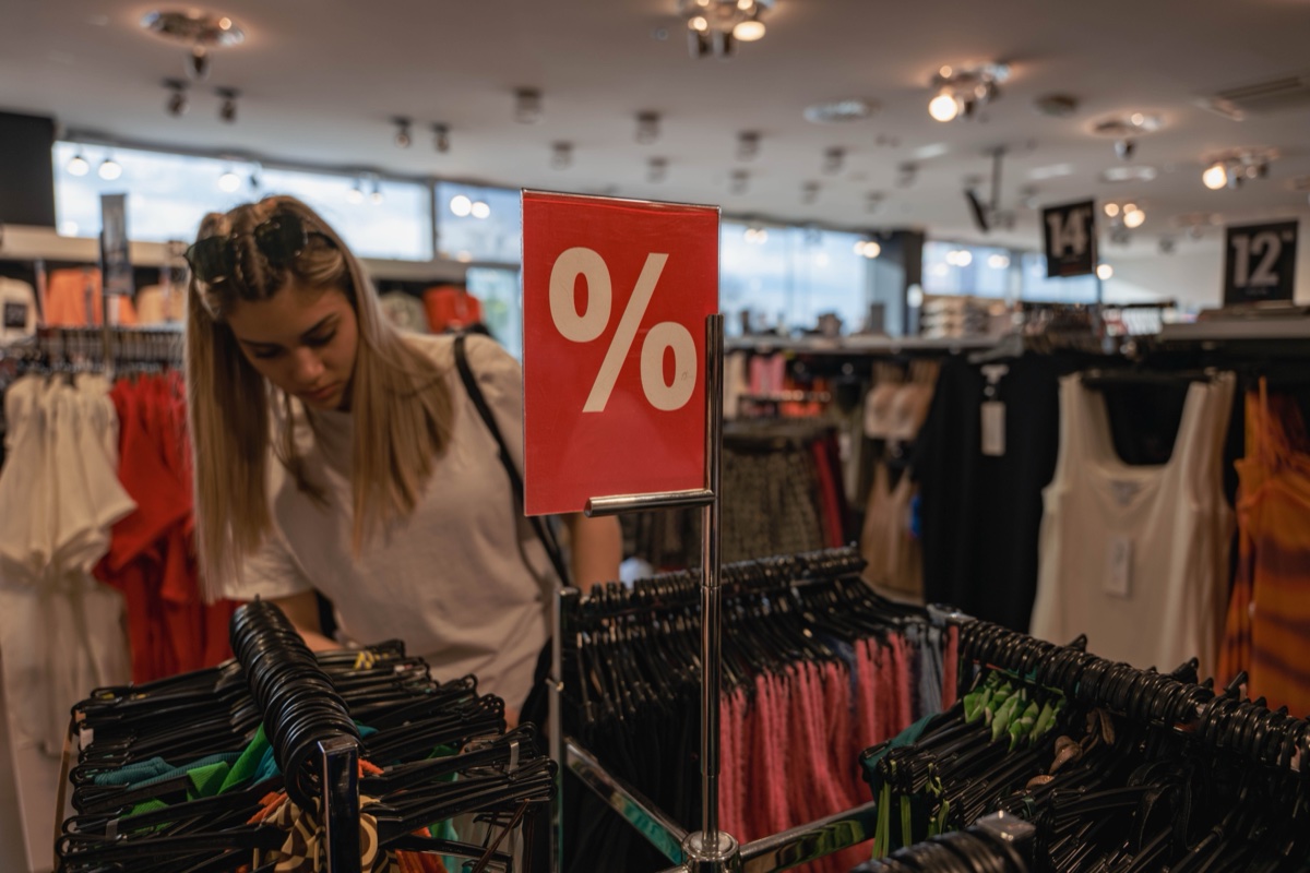 A red percent sign on a metal stand above a packed clothing rack while a shopper browses behind it
